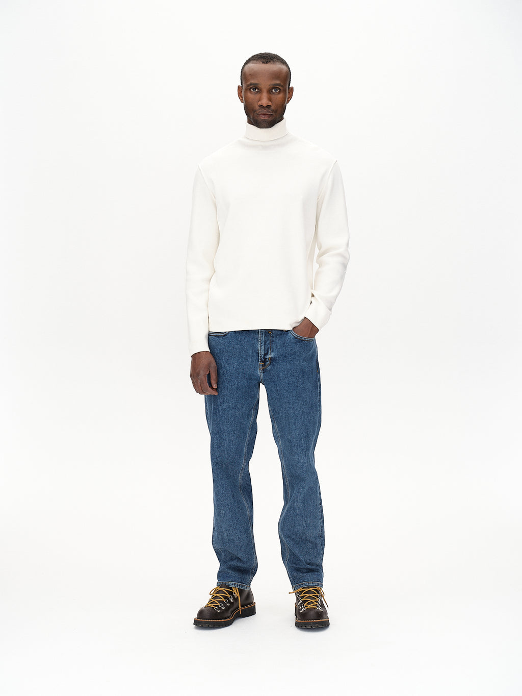 A man stands confidently against a plain white backdrop, embodying elegance with an edge. He dons a modern slim-fit white turtleneck, Max Cros - Mid Blue Denim jeans by Gabba, and dark brown boots with yellow laces. His left hand is casually in his pocket.