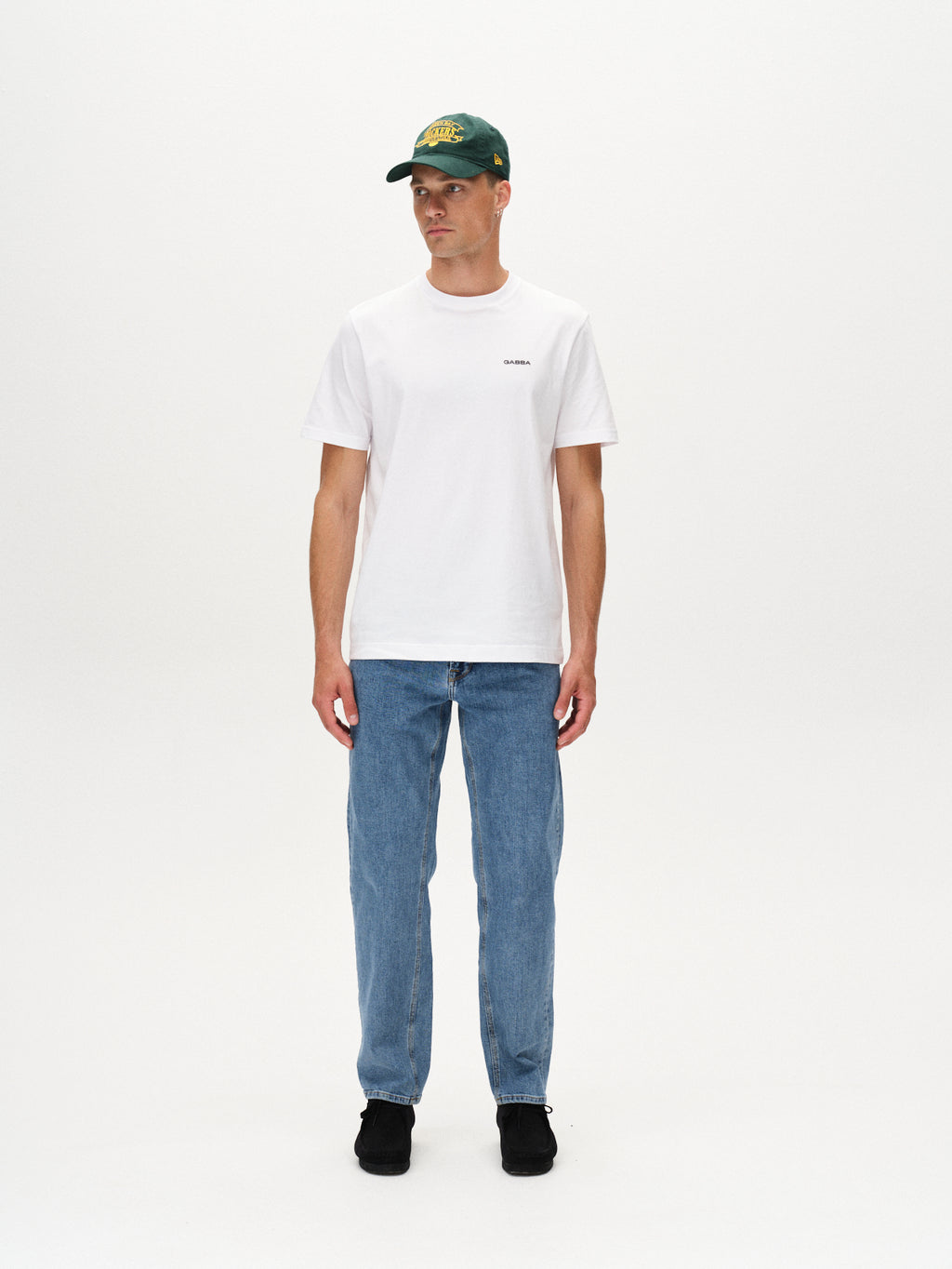 A person stands against a white background, wearing Gabbas Max Cros Lt. Blue Denim jeans, a white T-shirt, black shoes, and a green baseball cap. They look slightly to the side with arms relaxed at their sides.
