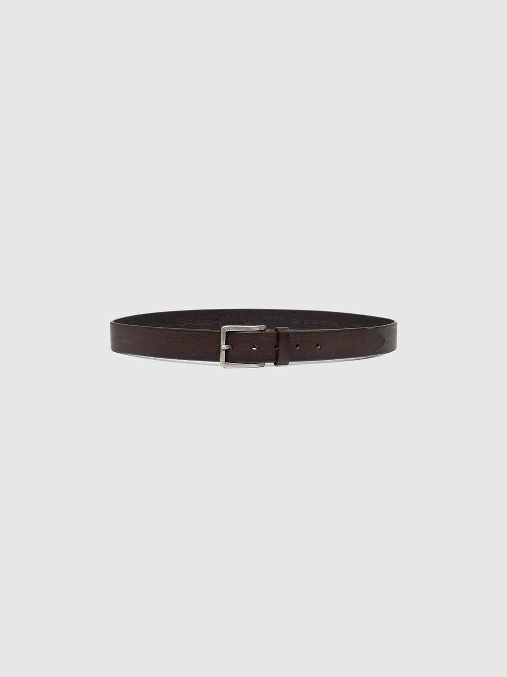 The Italy Belt - Brown by Gabba is a dark brown leather belt with a sleek metallic rectangular buckle, showcased on a plain gray background in a circular layout, highlighting its elegant and classic design.