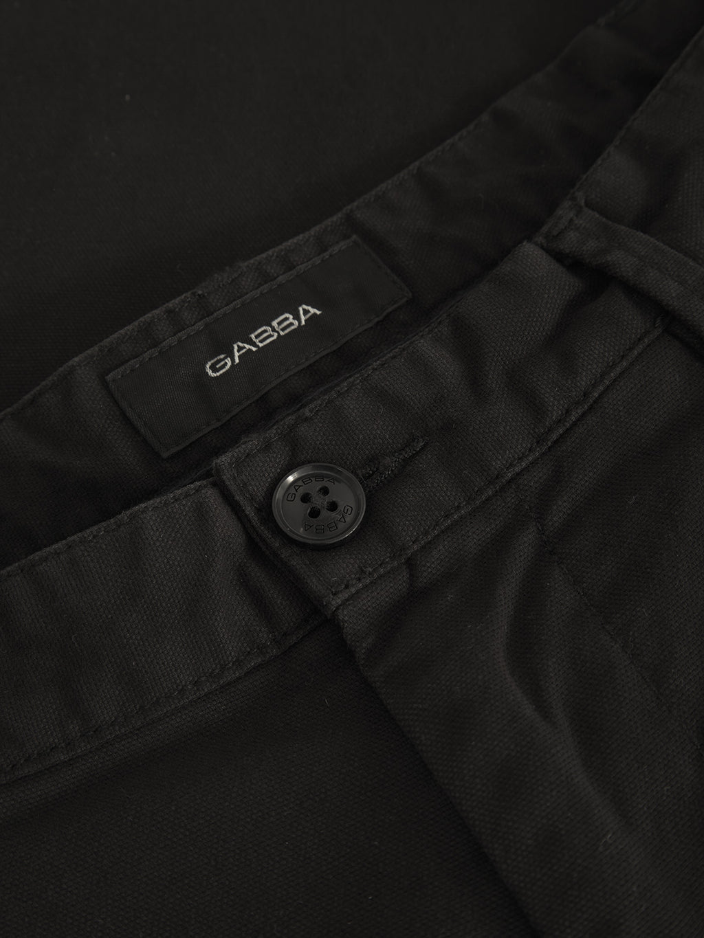 Close-up of Verona Ent - Meteorite black pants by Gabba, showcasing a buttoned waistband and sewn Gabba label. The durable textured fabric enhances its modern look, while the dark four-hole button adds detail. Soft lighting gently highlights this loose straight fit design.