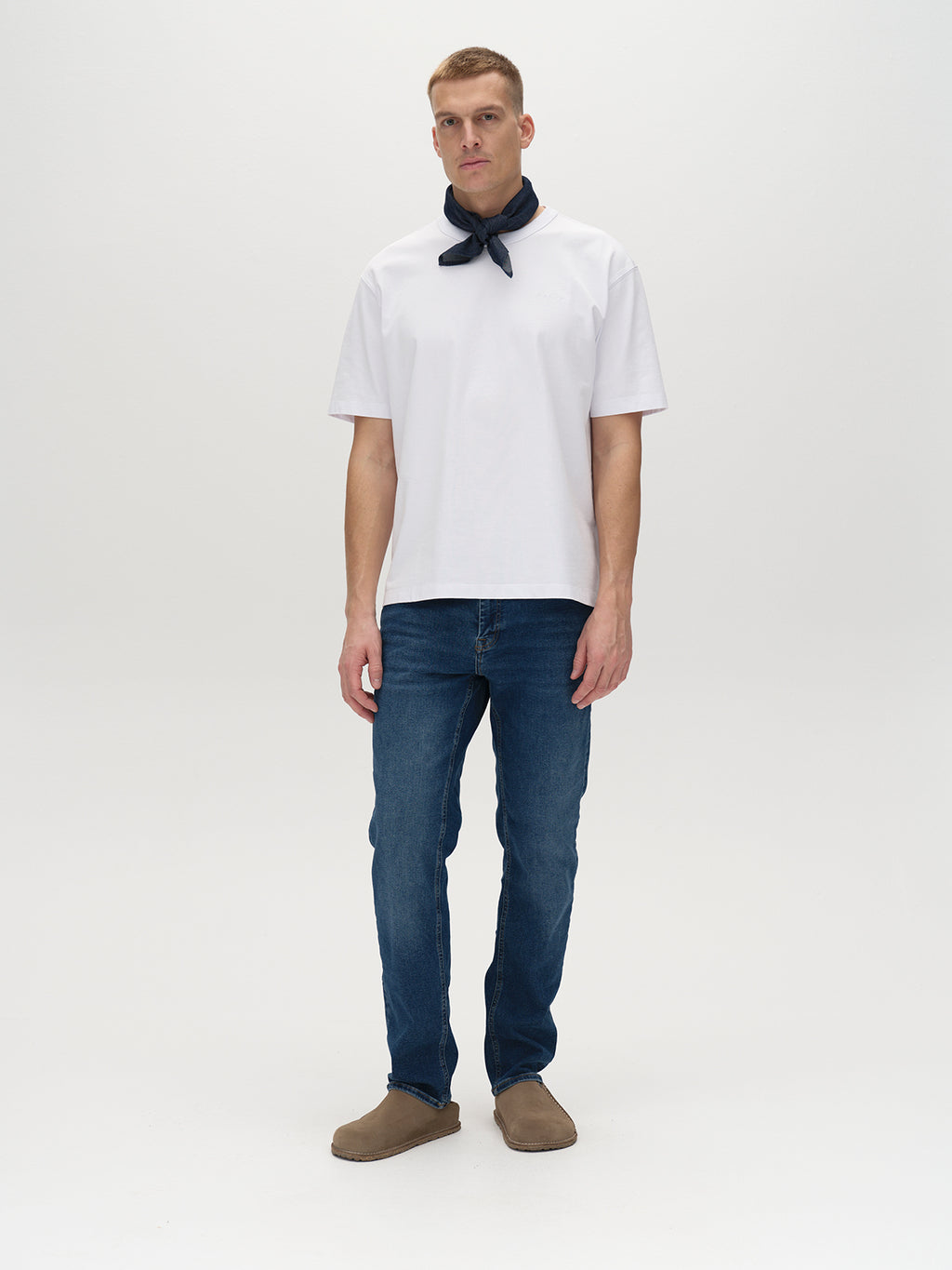 A person stands against a plain white background wearing a white T-shirt, Marc K4486 Limited - Mid Blue Denim jeans by Gabba, and brown slip-on shoes. They have a dark scarf tied around their neck, creating a classic look.