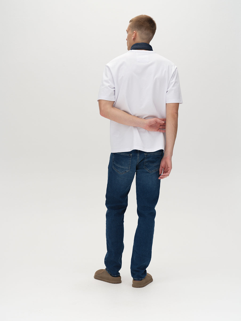 A person stands facing away in a white shirt with a dark collar, wearing Gabbas Marc K4486 Limited - Mid Blue Denim jeans and brown shoes on a plain white background. One hand is tucked behind their back, exuding timeless style.