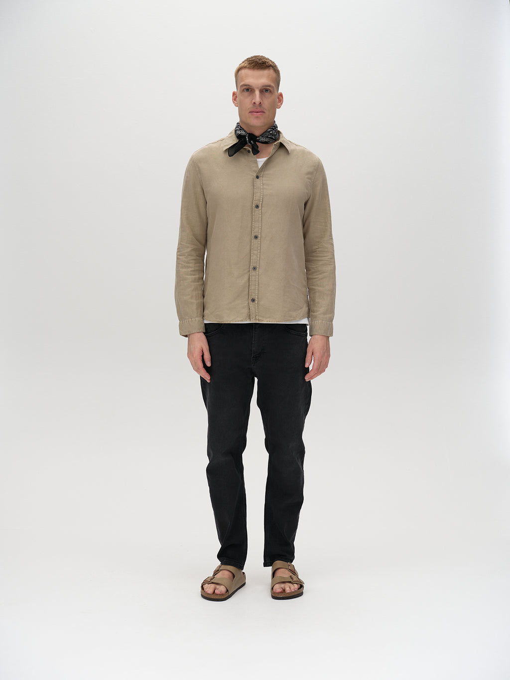 A man stands against a white background, wearing a beige shirt, Carl K4430 Limited - Grey Denim by Gabba, a black scarf around his neck, and brown sandals. He gazes directly at the camera with a neutral expression.