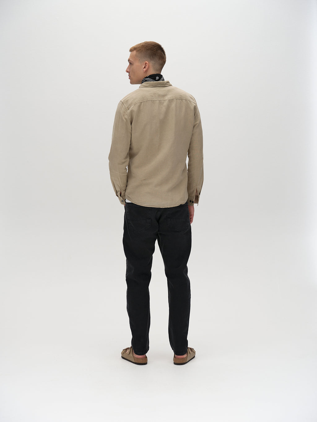 A person faces away from the camera, wearing a beige long-sleeve shirt and Gabbas Carl K4430 Limited - Grey Denim in a loose straight fit, complemented by sandals. The plain light background creates a minimalistic aesthetic.