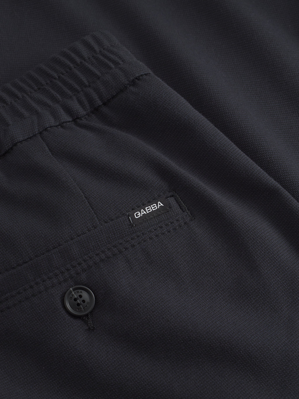 A close-up of navy mel fabric showcases an elastic waistband, a pocket with a small Gabba tag, and a sewn-on black button. The texture conveys classic style and comfort, reminiscent of the Napoli 19 Domo by Gabba with its subtly textured material.