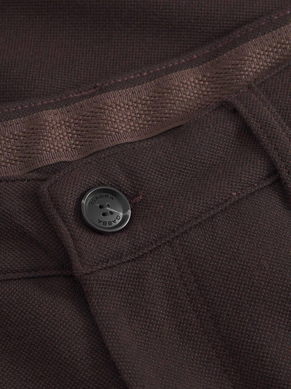 Close-up of Venza GKnit - Brown detail, featuring a black button engraved with Gabba on an intricately structured fabric. Impeccable stitching and texture highlight the craftsmanship, while its classic design hints at a regular straight-fitted pant style.