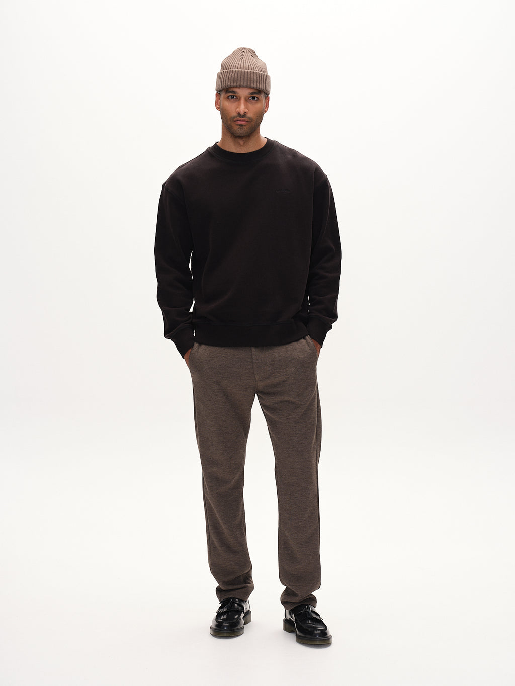 A person stands against a plain white background, wearing Gabbas Venza GKnit beanie in Sand. Theyre dressed in a dark sweatshirt, classic straight-fitted pants, and black shoes with their hands in their pockets.