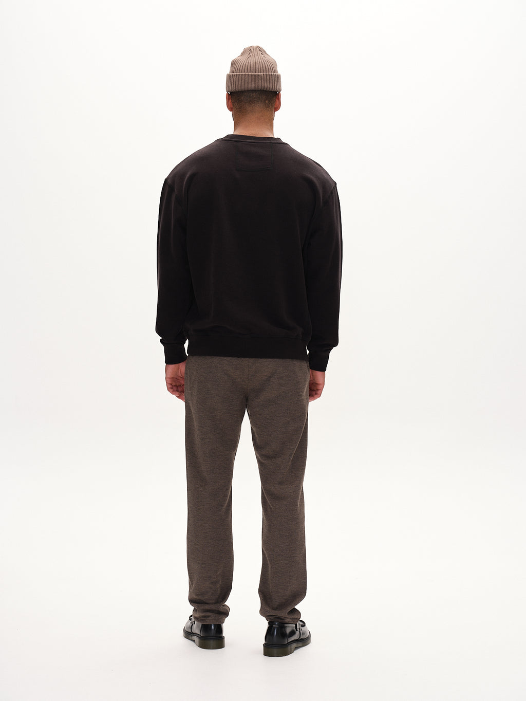 Wearing a classic black sweatshirt, straight-fitted pants, Gabbas Venza GKnit black shoes, and a beige beanie, a person stands facing away from the camera against a plain white background.