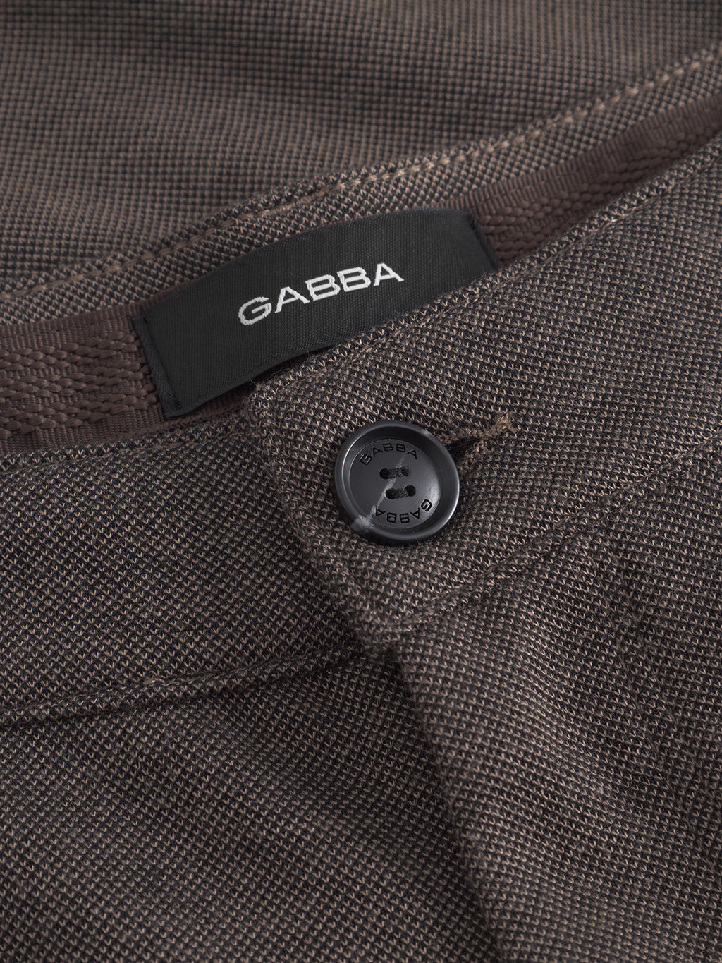 Close-up of Venza GKnit - Sand pants by Gabba, highlighting the straight-fitted, brown textured fabric with a black button. Focus is on the buttonhole area, showcasing the innovative weave and classic brand tag on the waistband.