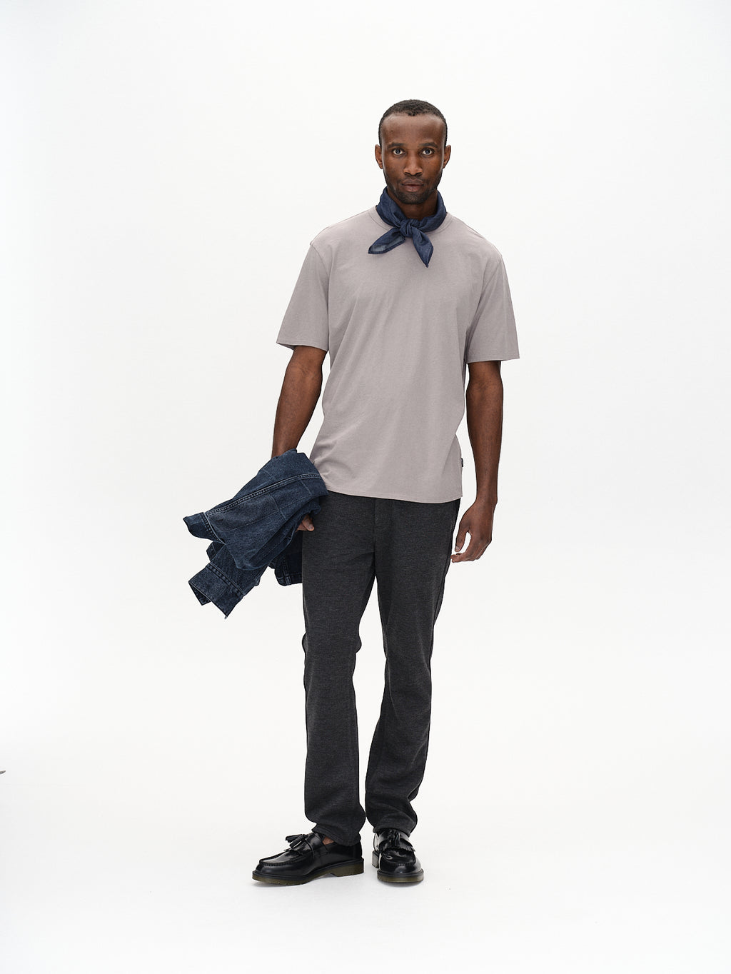 A man is set against a plain backdrop, wearing Gabbas Venza GKnit in Dk. Grey with classic straight-fitted pants and black shoes. He sports a dark blue bandana around his neck and holds a denim jacket made from innovative structure fabric in one hand.