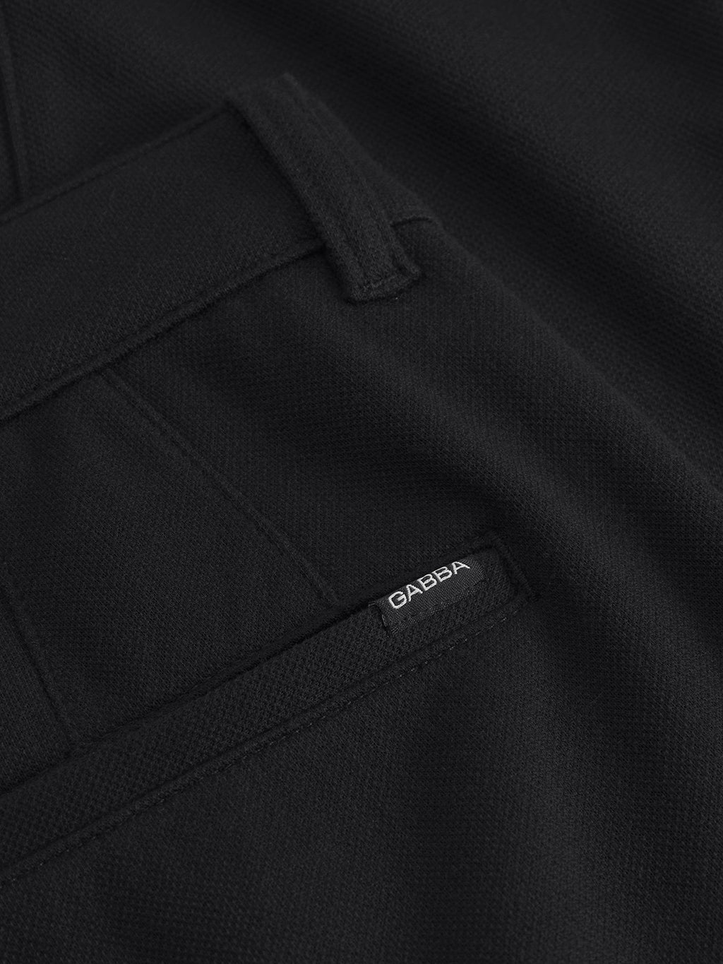 Close-up of black fabric from Gabbas Venza GKnit - Meteorite, highlighting detailed stitching and innovative structure. Part of a pocket features the classic rectangular GABBA label in white text. The texture implies the durability typical of straight-fitted pants.