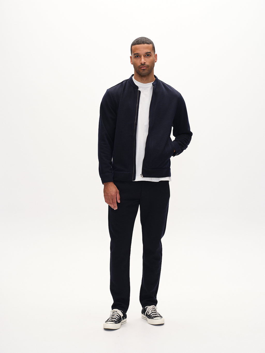 A man stands against a white background in an everyday look wearing Gabbas black zip-up jacket over a white T-shirt. Hes paired it with matching black pants and sleek black sneakers with white soles. His short hair adds to his neutral expression as he gazes at the camera.