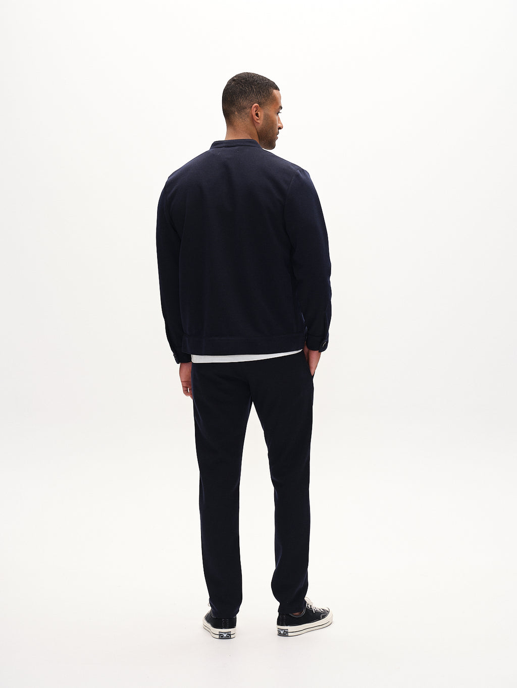 In a bright, minimalistic space, a person stands facing away in an everyday look, wearing a Gabba dark viscose mix jacket and pants, completed with Toto Gknit - Navy sneakers.