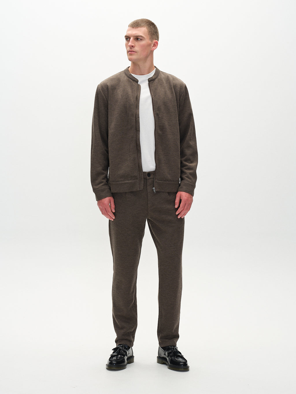 A person poses against a plain background in an everyday look, wearing the Gabba Toto Gknit - Sand zip-up jacket crafted from a comfy viscose mix, with matching pants and a white T-shirt underneath. They have short hair and black shoes, looking slightly to the side.