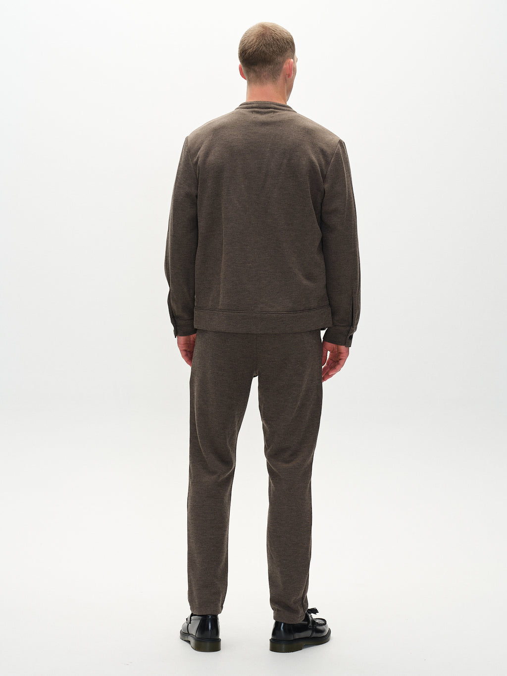 In a simple, well-lit studio, a man displays an everyday look in the Toto Gknit - Sand set from Gabba, crafted from a comfortable viscose mix. He stands facing away in his brown matching sweatshirt and pants, completing the ensemble with black shoes against a plain background.