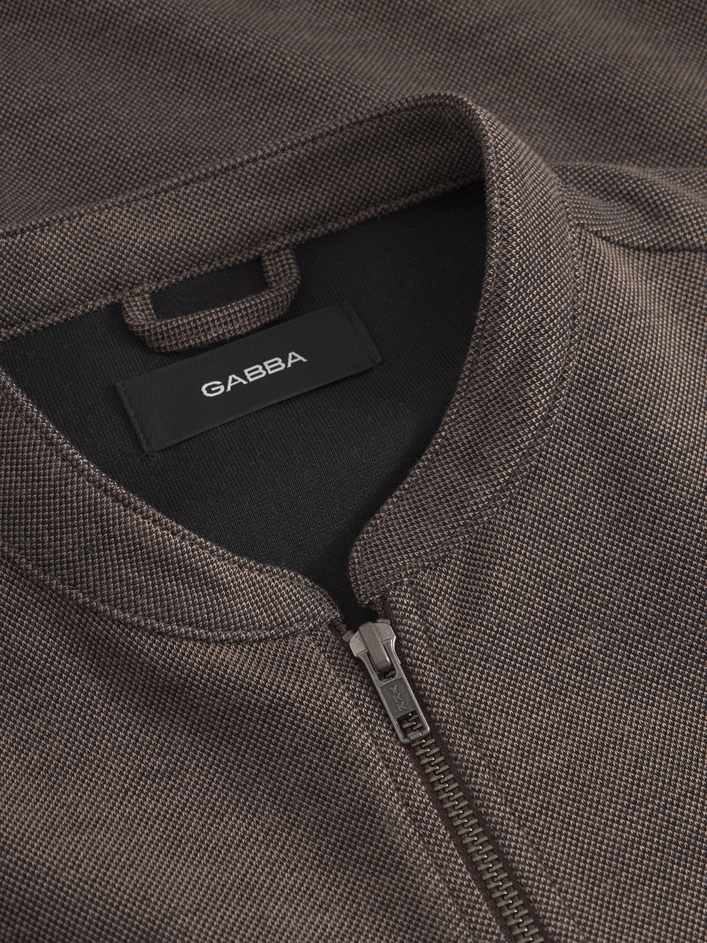 Close-up of the Toto Gknit - Sand by Gabba, a brown textured zip-up jacket made from viscose mix fabric with a dark collar and a visible black label with white text. The partially open zip reveals the black inner lining, perfect for an everyday look.