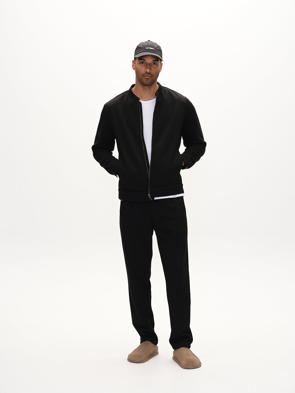 A man showcases an effortless everyday look against a plain backdrop, wearing Gabbas Toto Gknit - Meteorite: a dark jacket with sweat zip, matching pants, a white T-shirt, beige slip-ons, and a gray baseball cap. His hands are casually tucked into his pockets.