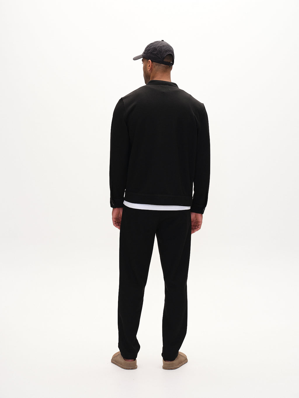 A man stands on a white background, wearing a black cap, sweat zip sweatshirt, and pants. He pairs his look with a white undershirt and brown Toto Gknit slippers by Gabba for an everyday style.