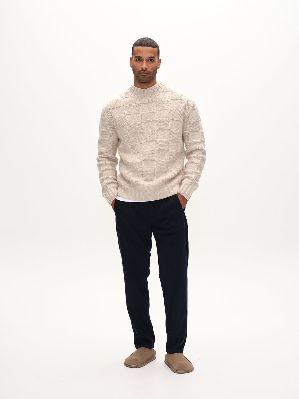 A man stands against a plain white background, wearing a beige textured sweater, Tito GKnit pants in navy by Gabba for comfort, and brown shoes. His hands are in his pockets as he looks directly at the camera.