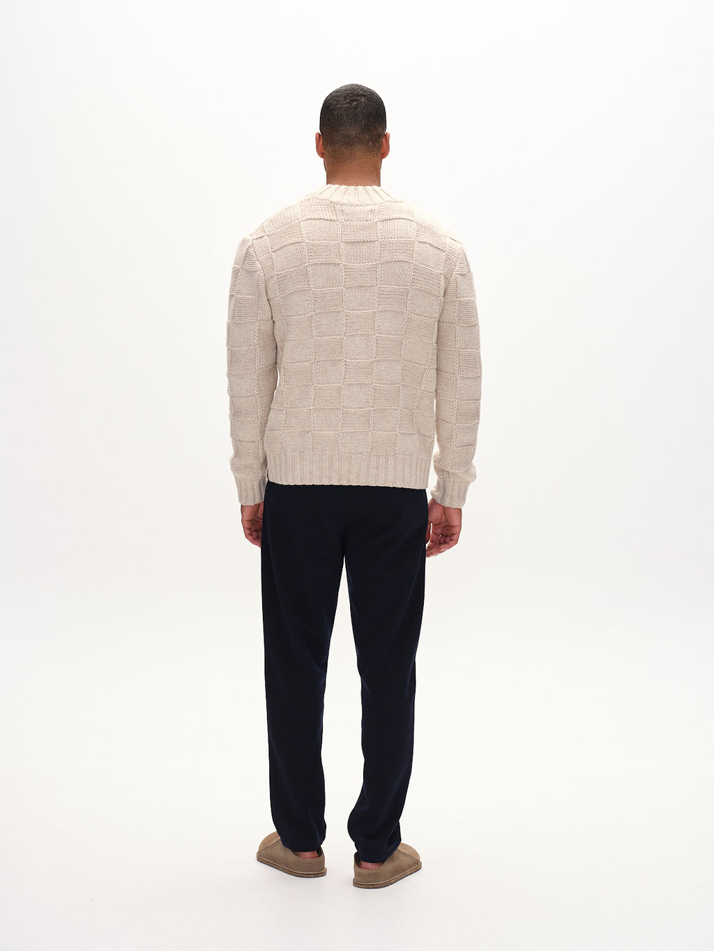 A man with short dark hair faces away, wearing an intricately knit cream sweater, paired with Gabbas navy Tito GKnit pants and brown slippers. The plain light gray background enhances the comfort of his outfit.