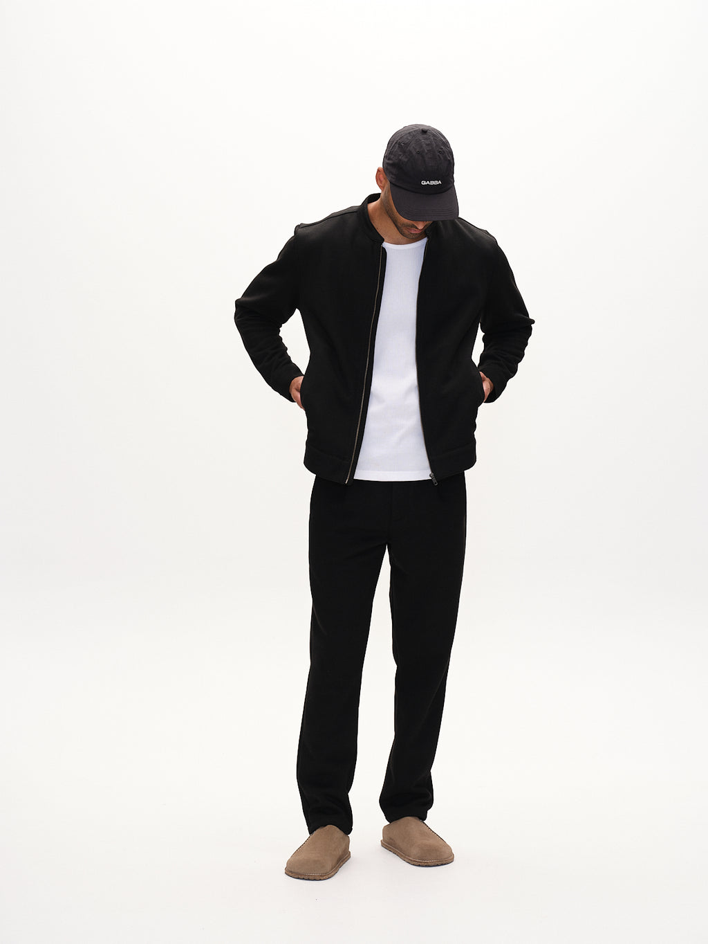A person stands against a plain white background wearing a black cap, black jacket, white shirt, and beige shoes. Completing their stylish look are Gabbas Tito GKnit - Meteorite pants. Hands tucked in pockets, they look down with confidence in these sleek, regular straight-fit knit pants.