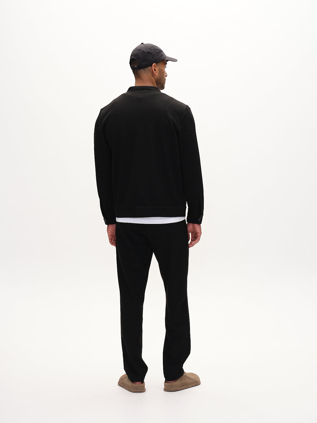 A person in a Gabba Tito GKnit - Meteorite pants, regular straight fit, with a white shirt, black jacket, gray cap, and brown shoes stands against a plain white background showcasing the innovative knit structure.
