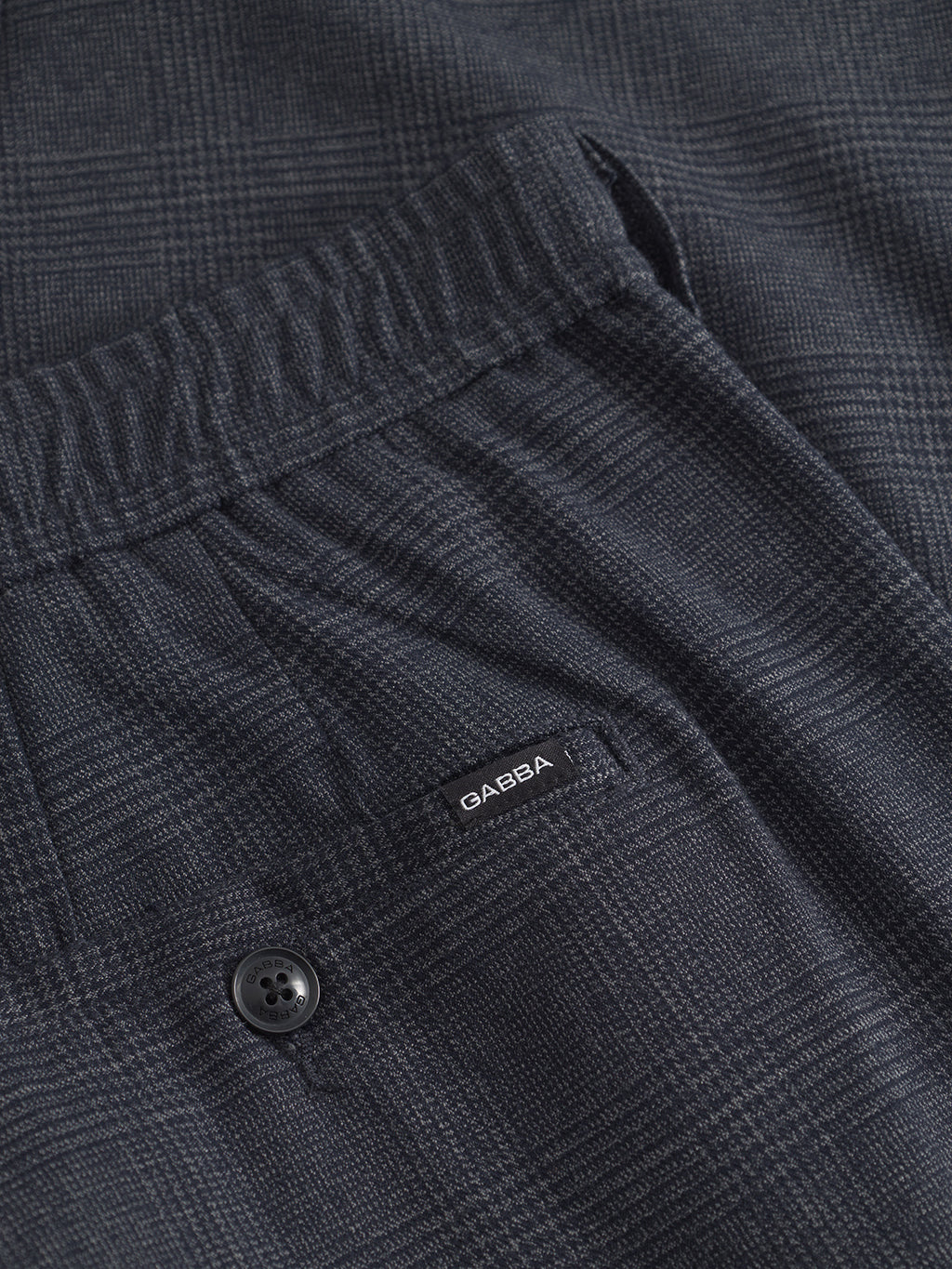 Close-up of navy Napoli 19 Field Pants by Gabba, featuring a dark gray plaid pattern, elastic waistband, small rear pocket with a visible label, and button below the pocket for classic style and comfort.
