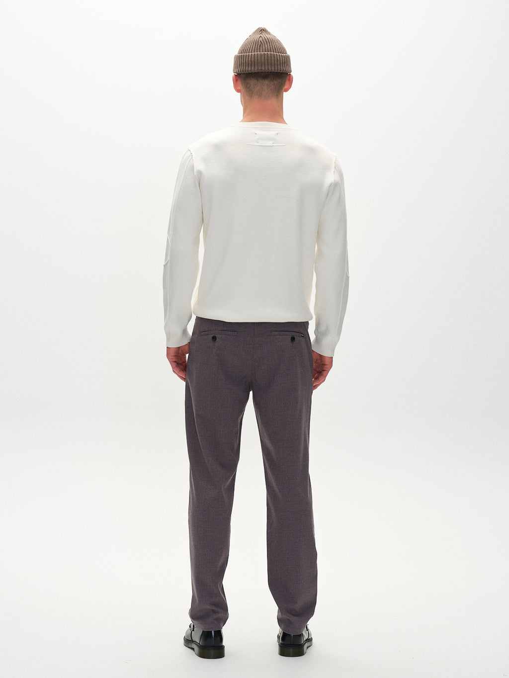 Facing away, the person wears a beige beanie, white sweater, Gabbas Napoli 19 Field Pants in grey, and black shoes. The plain white background highlights their comfortable and casual style.