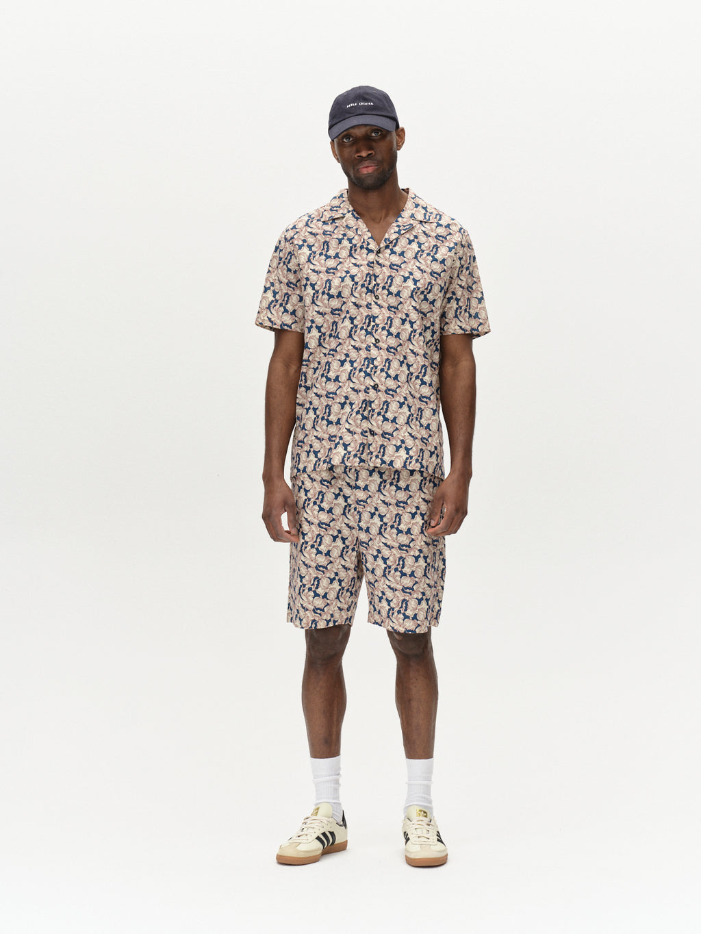 A man wears the Gabba Sven Reg Resort Epo SS - Blue Pattern shirt and shorts set, exuding a retro flowers vibe. He pairs it with a black baseball cap, white socks, and beige sneakers with black stripes, standing relaxed against a plain white background.