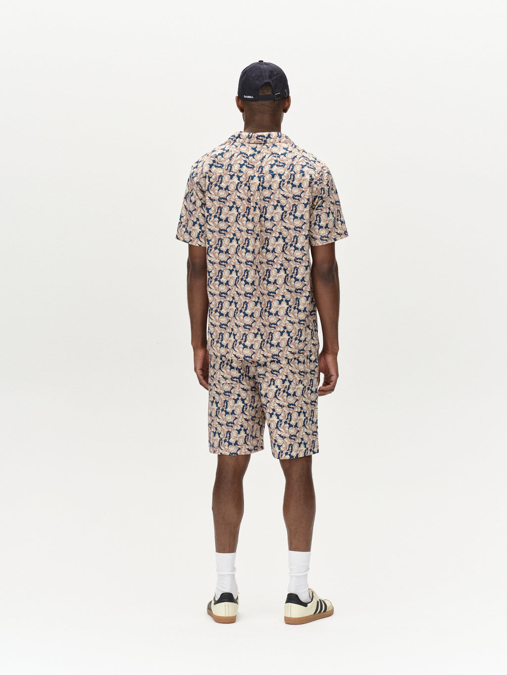 Facing away in a Gabba Sven Reg Resort Epo SS shirt, the person showcases a premium outfit with abstract blue and beige patterns reminiscent of retro flowers, paired with white socks and Adidas sneakers. Their look is completed by a black baseball cap against the plain white backdrop.