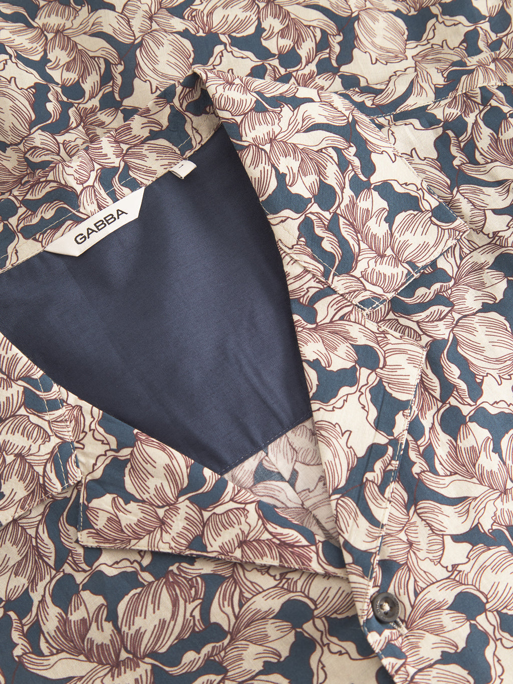 A close-up of the Gabba Sven Reg Resort Epo SS jacket reveals a retro blue and beige floral pattern, with a partially open design showcasing its intricate craftsmanship. The navy lining holds a visible Gabba tag. Made from 100% cotton, it highlights exquisite detail in every stitch.