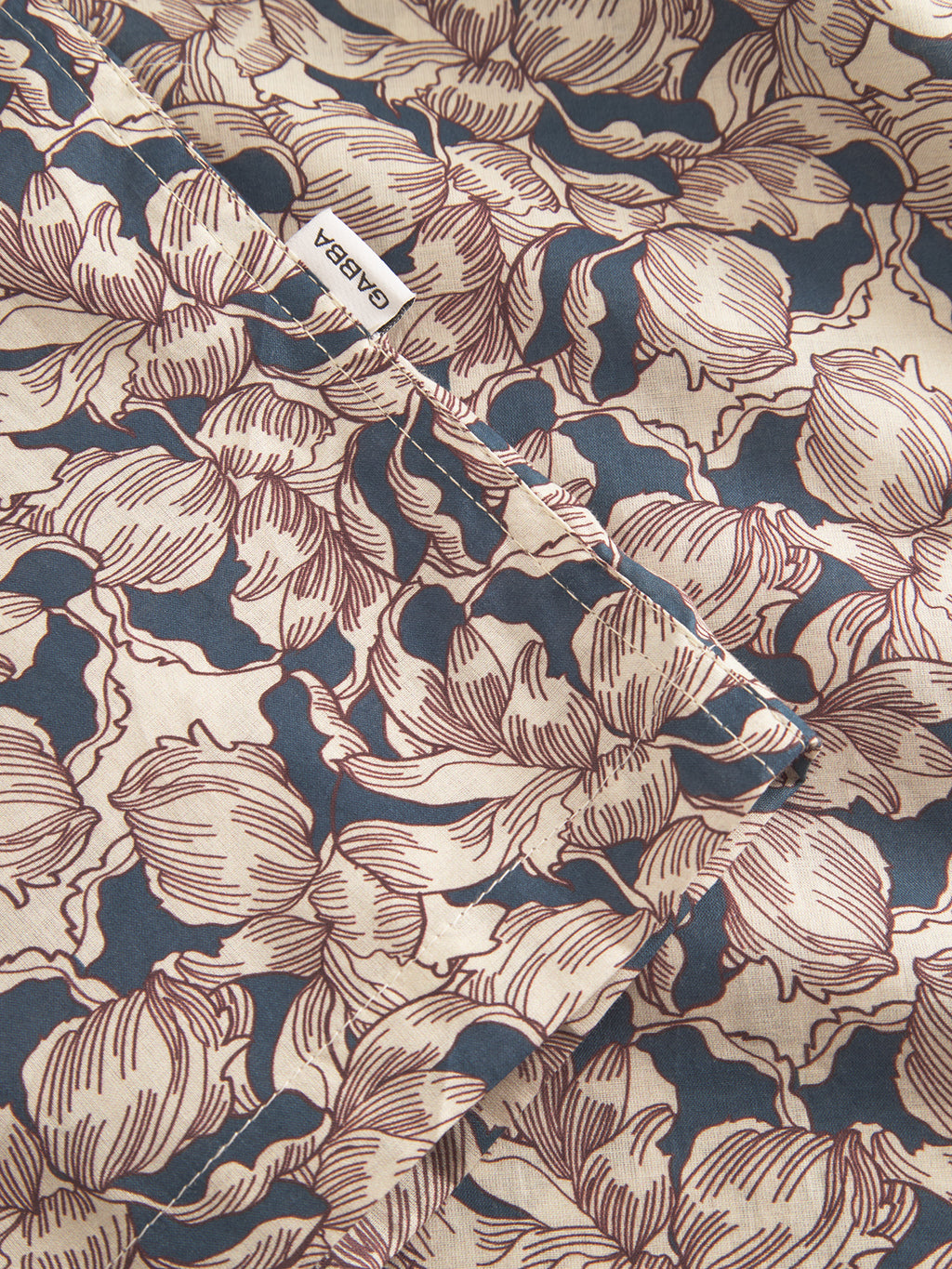A close-up of a Gabba Sven Reg Resort Epo SS shirt fabric in retro beige, navy, and brown floral, made from 100% cotton with stylized petals. A small white ARABA label is sewn on.