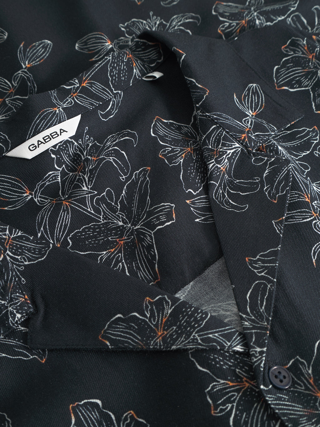 A close-up of the Sven Reg Resort Lofi SS - Flower Mix by Gabba showcases a black fabric with white floral patterns and orange accents. The design, complete with a tag labeled Gabba and a black button, exudes a vacation-ready vibe, enhanced by intricate outlines of flowers and leaves.