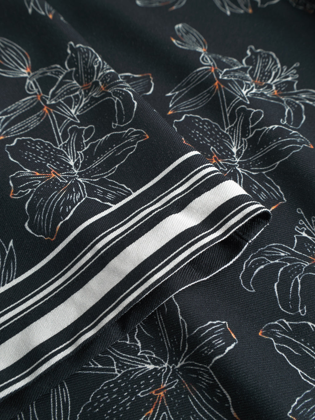 A close-up of Gabbas Sven Reg Resort Lofi SS - Flower Mix reveals black fabric with a white floral pattern and orange accents reminiscent of tropical lilies. The folded edge has parallel white and black stripes, perfect for resort-inspired shirts.