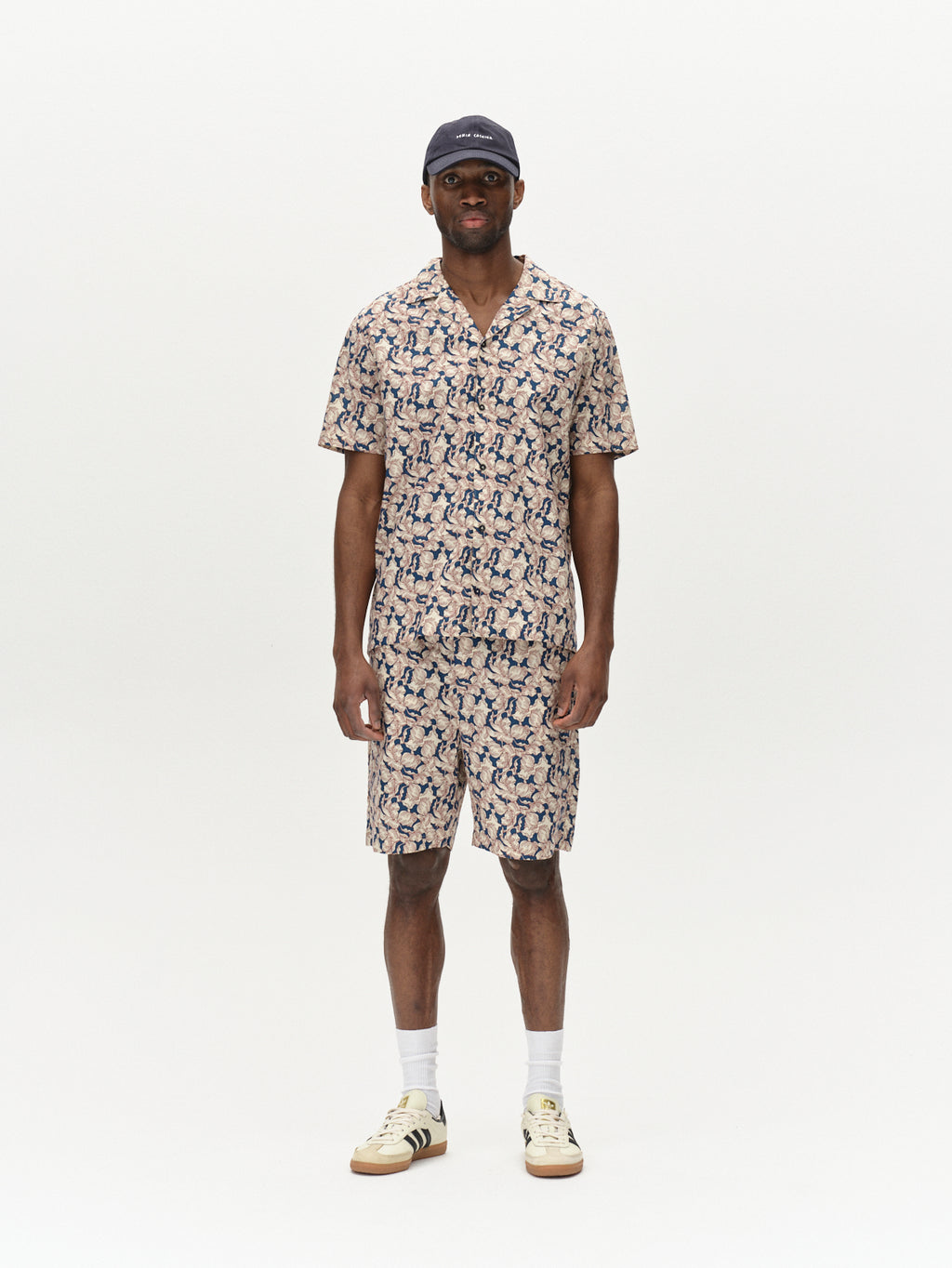 Against a plain white background, someone models the Porter Epo - Blue Pattern set from Gabba, featuring premium shorts and a shirt. A dark cap, white socks, and light-colored sneakers with dark stripes enhance this summer-inspired look that effortlessly combines comfort and style.