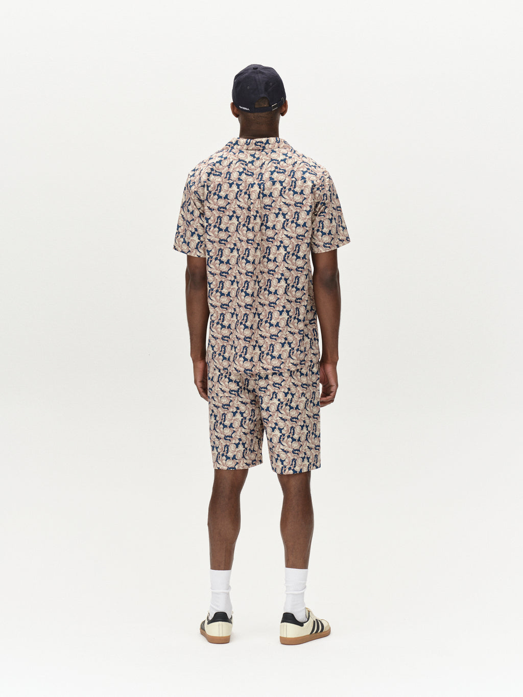 A person faces away wearing Gabbas Porter Epo ensemble, featuring a short-sleeve shirt and premium shorts with matching abstract blue patterns. Made from 100% cotton, they accessorize with a black cap, white socks, and striped sneakers on a plain white background.