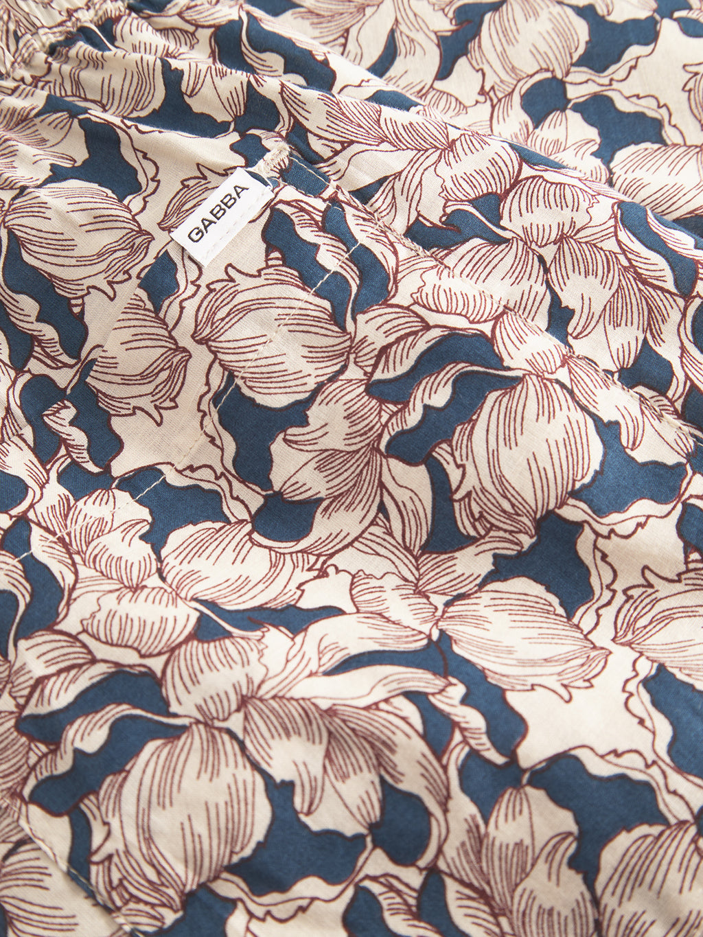 The Porter Epo - Blue Pattern by Gabba is a summer-inspired fabric in calming beige and blue floral hues. Made from 100% cotton, it includes a small white label featuring Gabba, ideal for crafting premium shorts.