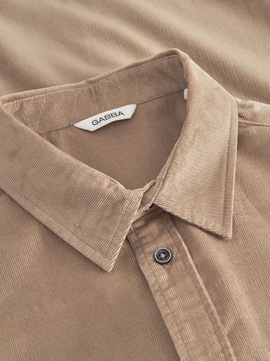 Close-up of the Sven Reg Cord SS shirt in sand greige by Gabba, featuring a classic collar, partially buttoned front, and a textured ribbed appearance. The collar tag reveals its 100% cotton construction.