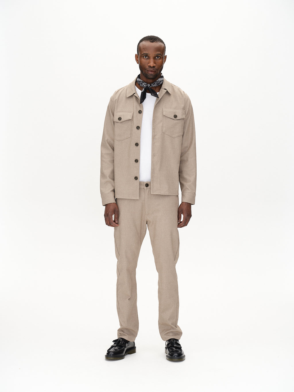 A man stands against a white background, wearing a beige button-up jacket and matching pants. His black and white scarf reveals the Clipper Shafi - Brown shirt by Gabba underneath, adding flair to his outfit. The ensemble is complete with black shoes that feature elegant stitching and a refined wool-like look.