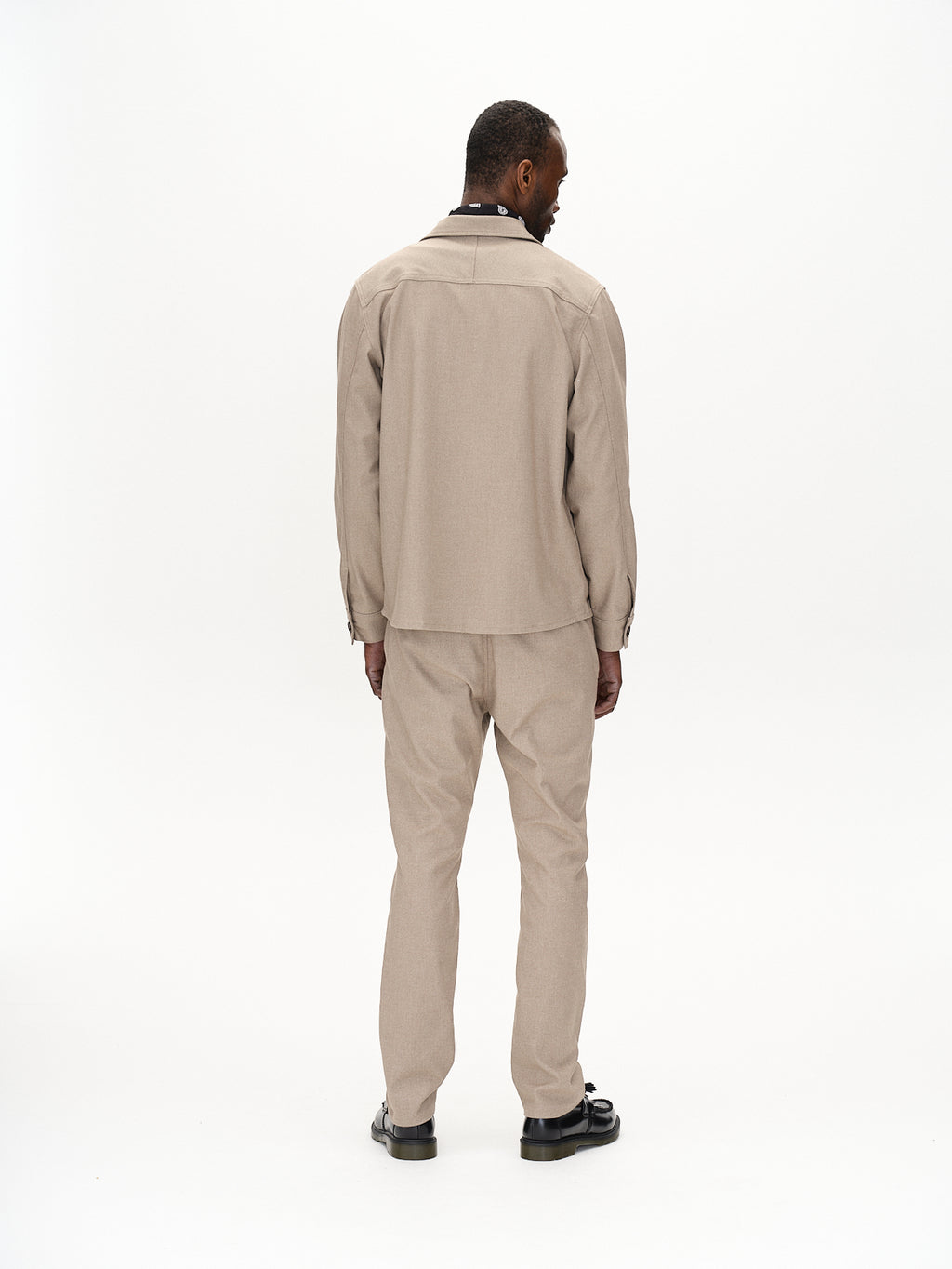 A man stands facing away in a Gabba Clipper Shafi shirt, exuding a wool-like appearance. The brown long-sleeve top and matching pants feature elegant stitching, with his short hair complementing the minimalist white space around him.