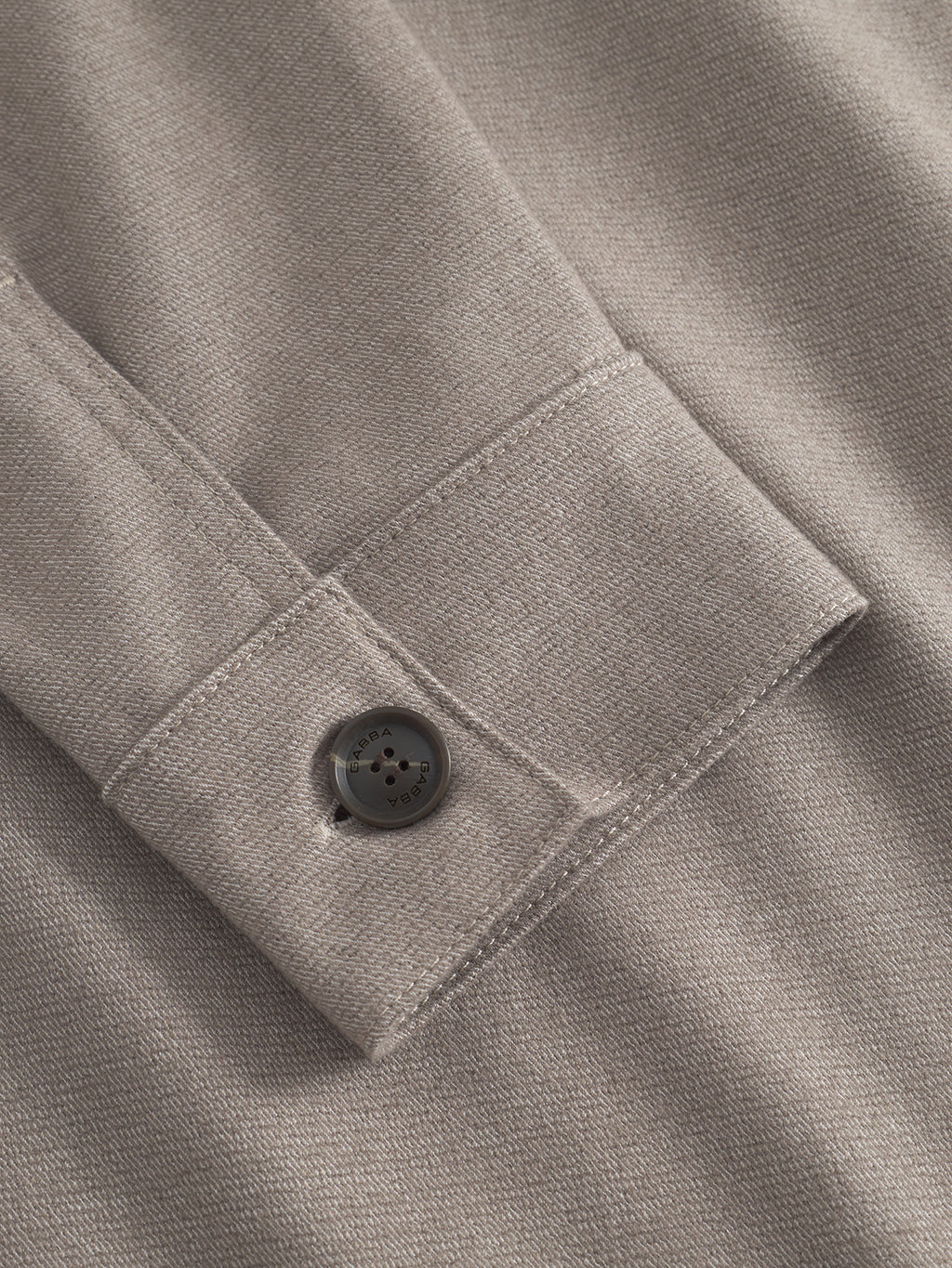 Close-up of gray fabric with a shiny button on a folded edge, highlighting elegant stitching akin to the Gabba Clipper Shafi – Brown shirt. The texture reveals a fine weave and wool-like look, adding subtle sheen to the material.