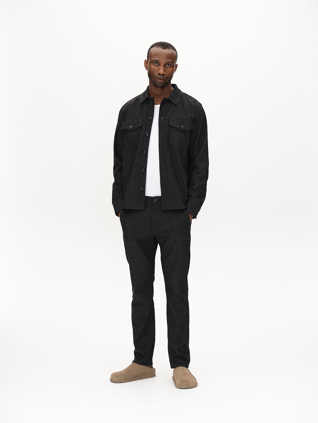 Wearing a dark jacket and matching pants, the person pairs them with a crisp Gabba Clipper Shafi - Black shirt featuring dual chest pockets and beige slip-on shoes. Standing against a white background, they present a wool-like appearance with a neutral expression, hands casually by their sides.