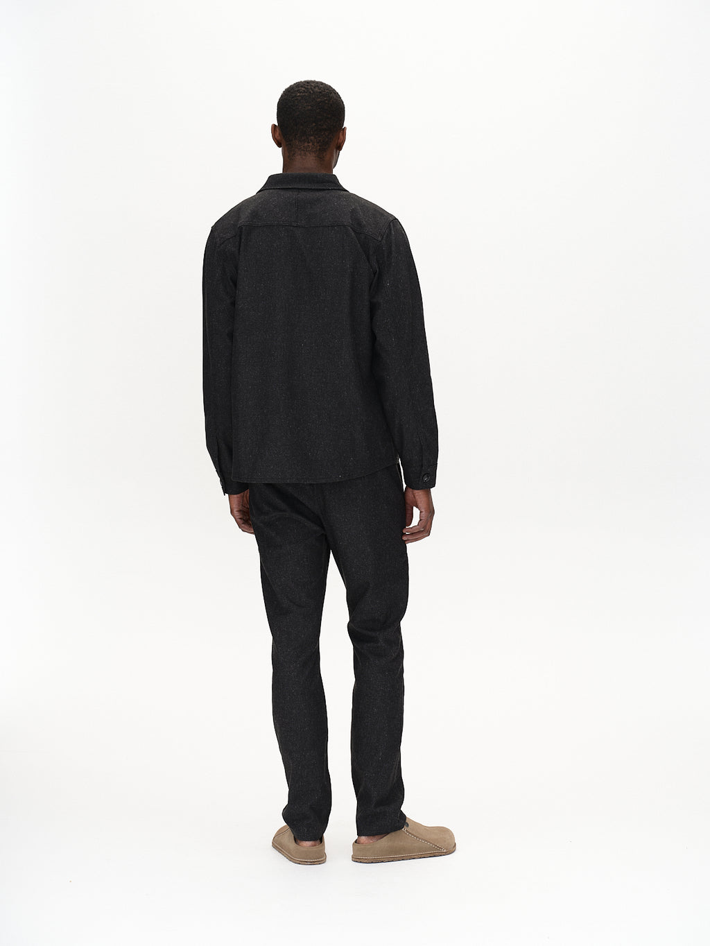 Against a white backdrop, an individual faces away wearing Gabbas Clipper Shafi shirt in black with dual chest pockets, matching pants, and tan shoes that have a wool-like texture.