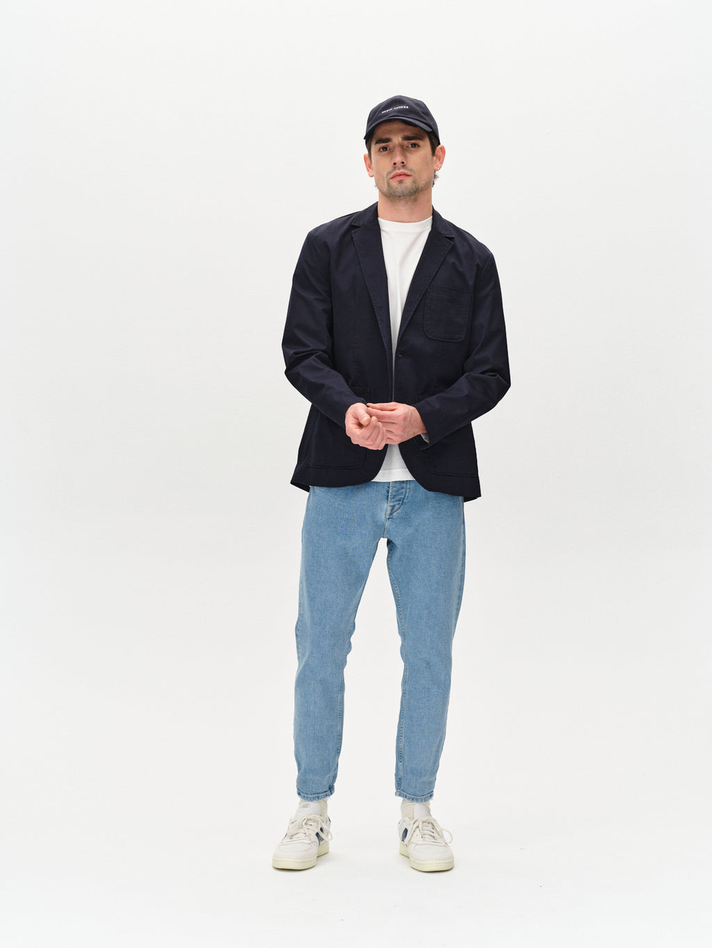 A man poses against a white background in easy style, wearing Gabba’s Alex Cross Lt. Blue Denim jeans with a relaxed crotch, paired with a black cap and jacket, white t-shirt, and white sneakers, hands clasped in front.