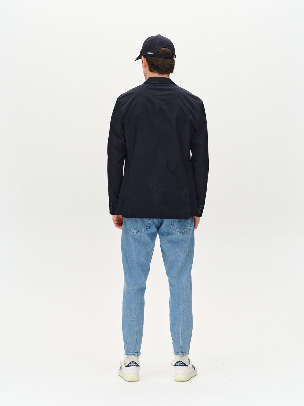 A person stands facing away from the camera, wearing Gabba’s Alex Cross - Lt. Blue Denim jeans with a relaxed crotch, navy cap, dark navy jacket, and white sneakers against a plain white background—showcasing effortless style.