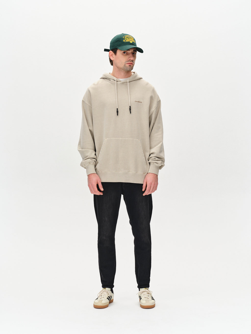 A person stands against a plain white background in a trend-focused look: a beige hoodie, Gabba Alex Telsa Black Denim jeans with relaxed crotch, white sneakers with gum sole, and a green baseball cap featuring yellow lettering.