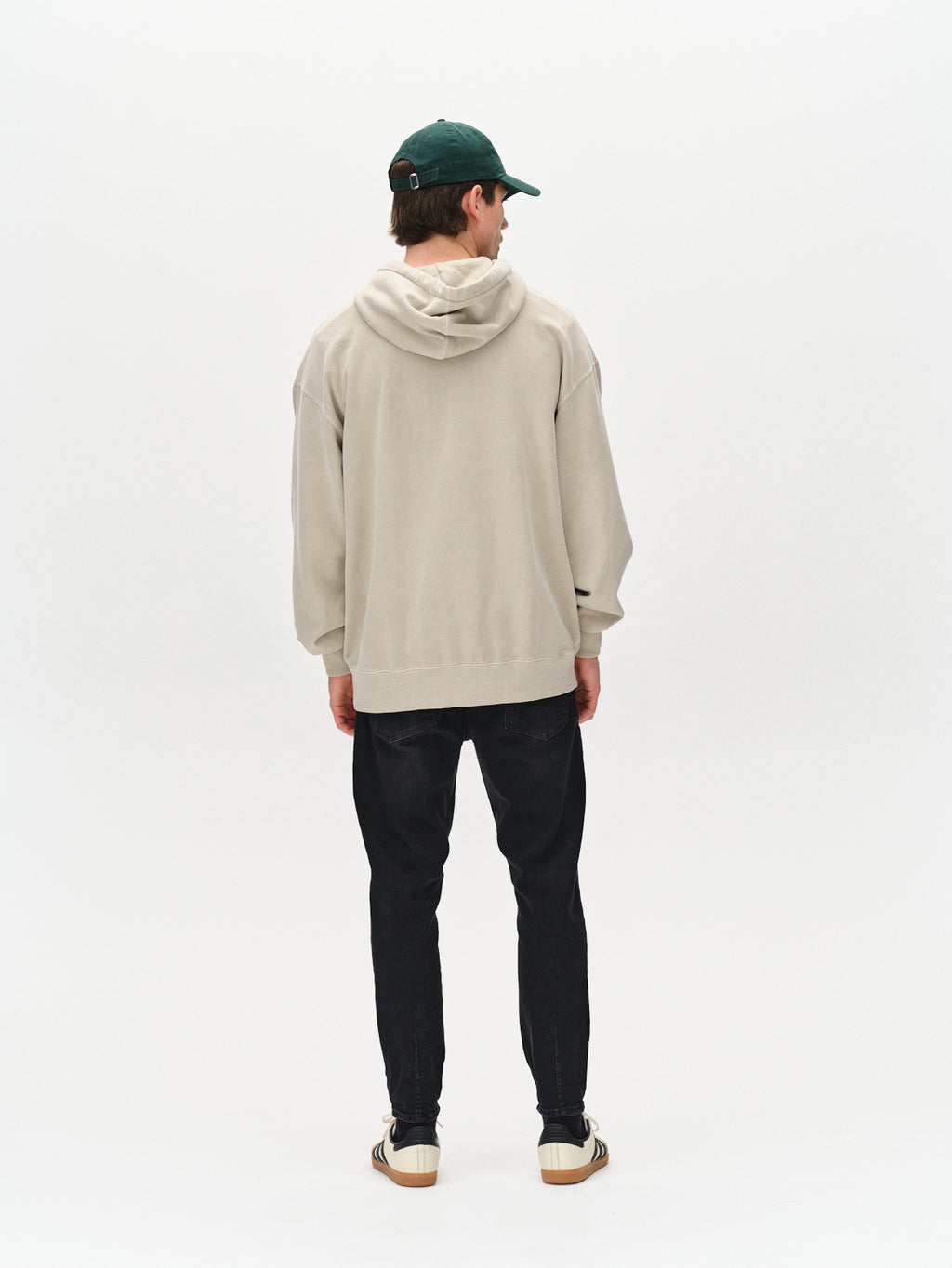 A person faces away against a white background, wearing a beige hoodie, Gabba Alex Telsa - Black Denim jeans, white striped sneakers, and a dark green baseball cap.