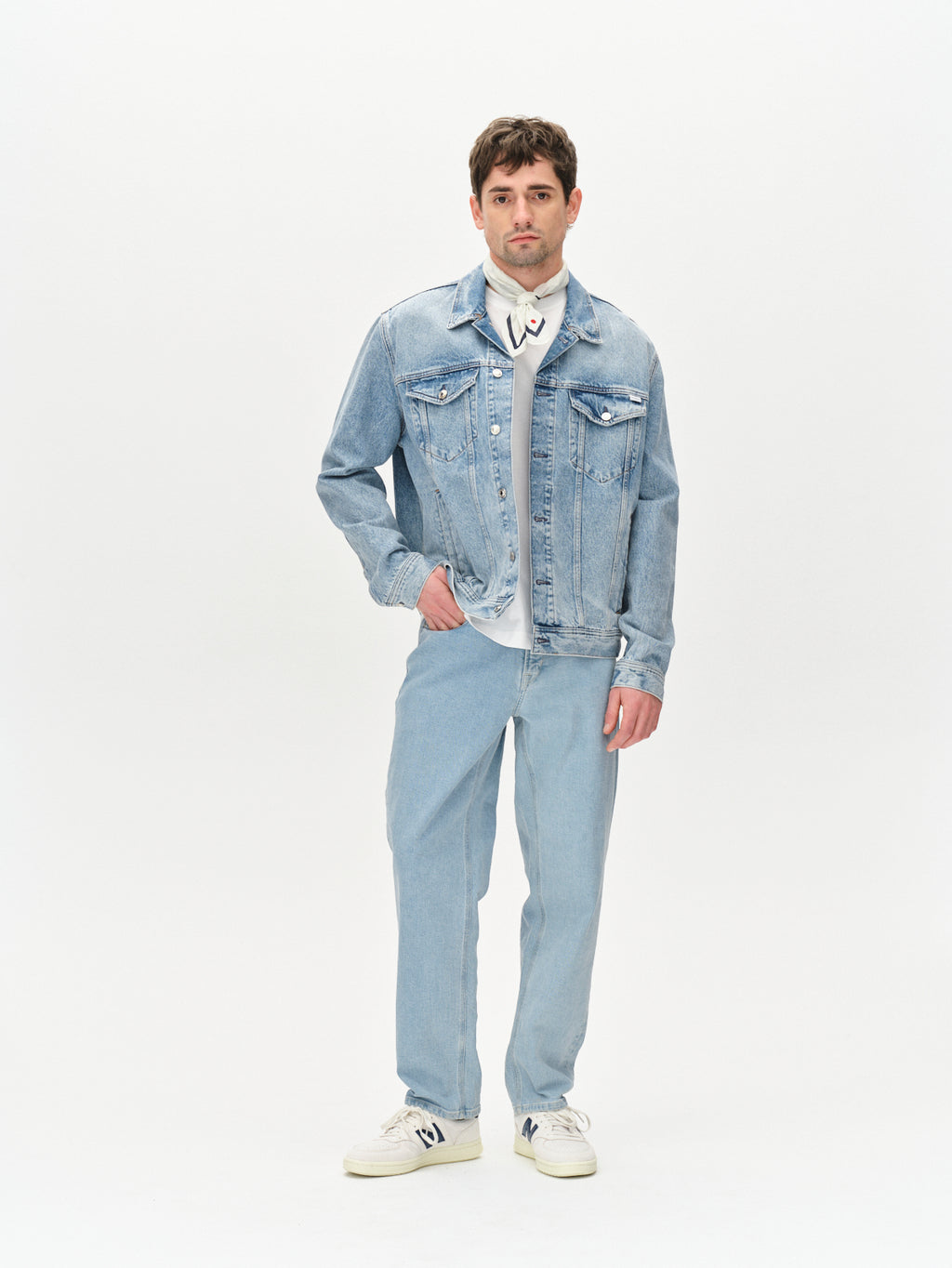 A man stands against a white background wearing the Gabba Max Cross - Lt. Blue Denim jacket and jeans, paired with a white shirt and sneakers. His modern outfit is completed by short brown hair and a neutral expression.