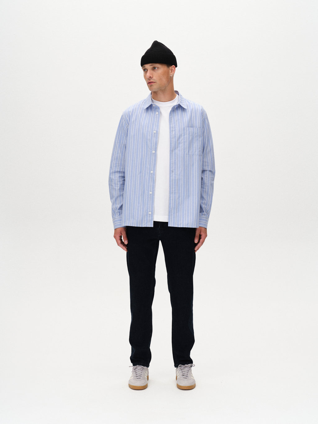 A man models a modern classic look in Gabba’s Marc Panter - Dark Blue Denim jeans, styled with a black beanie, light blue striped shirt over a white tee, and gray sneakers against a plain white background.