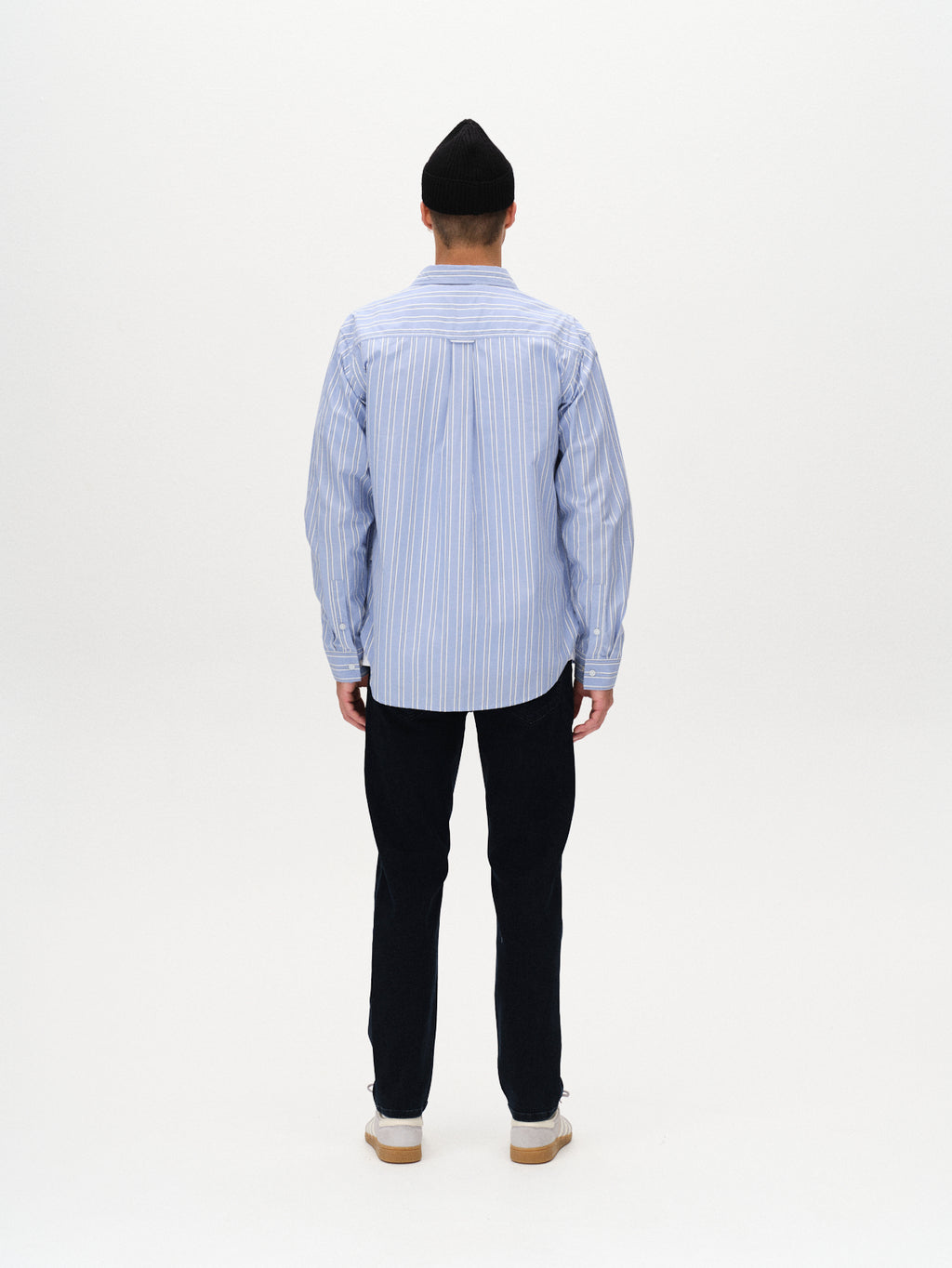 A person wearing a black beanie, light blue striped button-up shirt, Gabba Marc Panter dark blue denim jeans with a slim thigh fit, and white shoes stands facing away against a plain white background for a modern classic look.